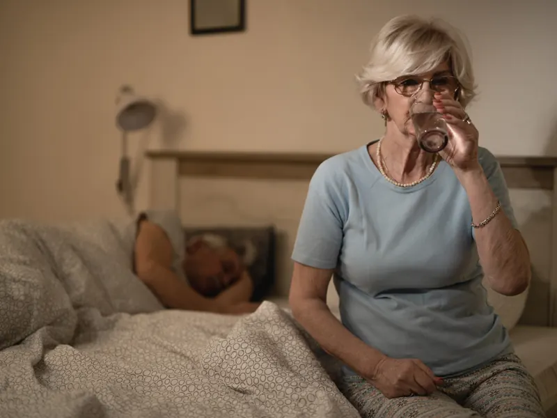 Idosa acordada à noite bebendo água enquanto o marido dorme